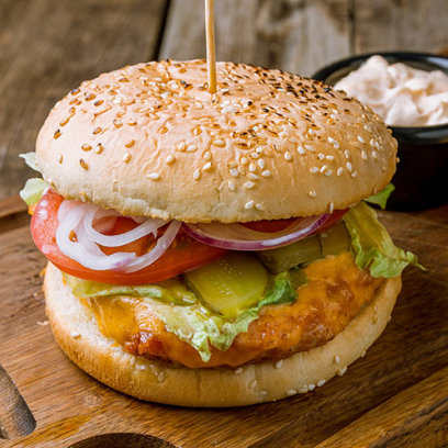 Um hambúrguer com um bolinho de peixe, alface, tomate, cebola e picles em um pão com gergelim, servido em uma tábua de madeira com uma tigela pequena de maionese ao lado.