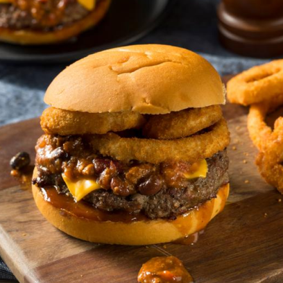 A imagem mostra um hambúrguer apetitoso em um pão com gergelim. O hambúrguer é recheado com uma generosa porção de carne grelhada, queijo derretido, alface fresca, fatias de tomate e cebola roxa. Ao lado do hambúrguer há uma porção de batatas fritas douradas e crocantes. O conjunto está apresentado em uma tábua de madeira rústica. Há um espeto decorativo inserido no centro do hambúrguer.